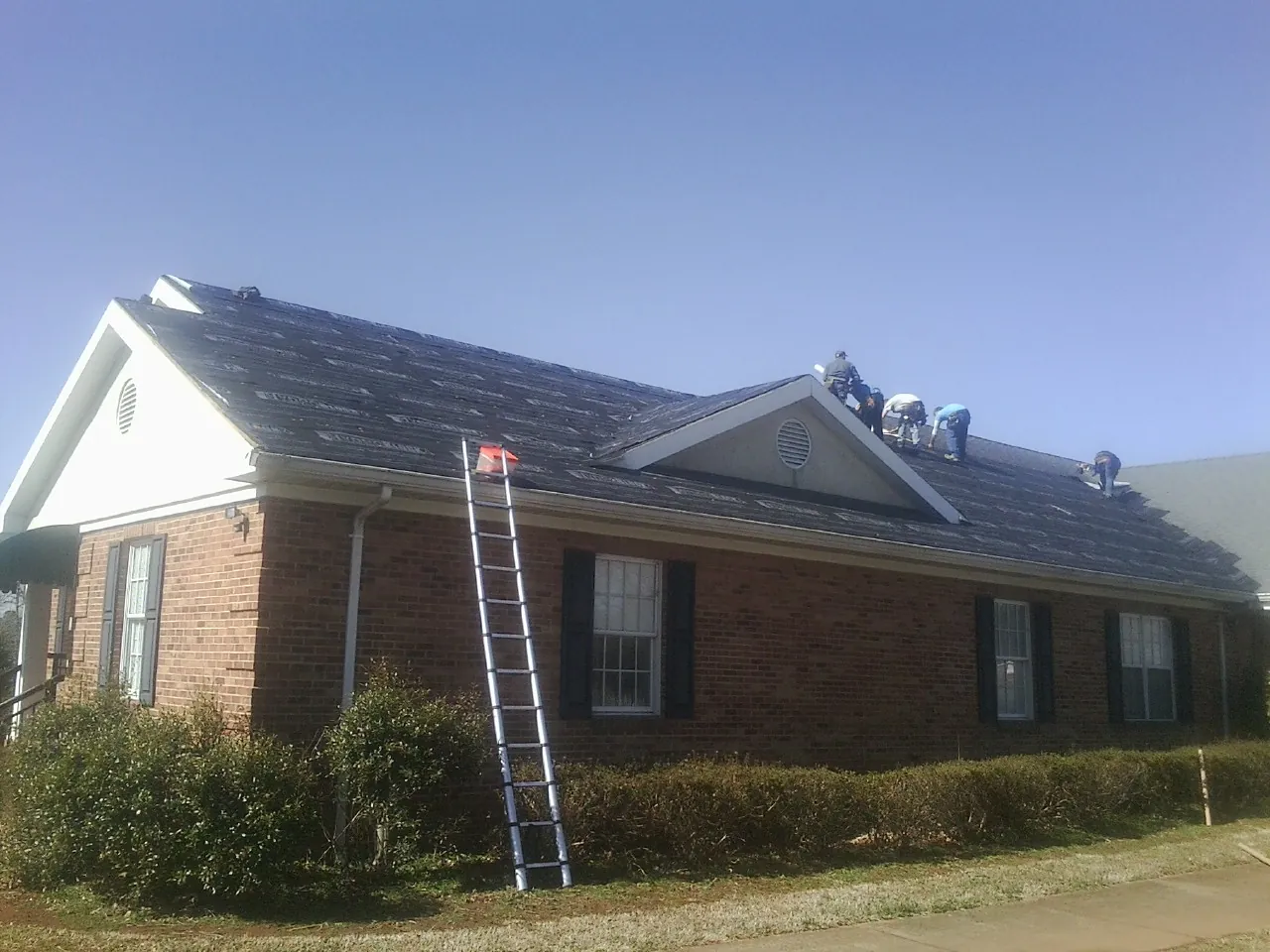 Crew tearing shingles off a brick ranch roof with a ladder set — Emergency Roof Repair work by our crew in Cave Springs