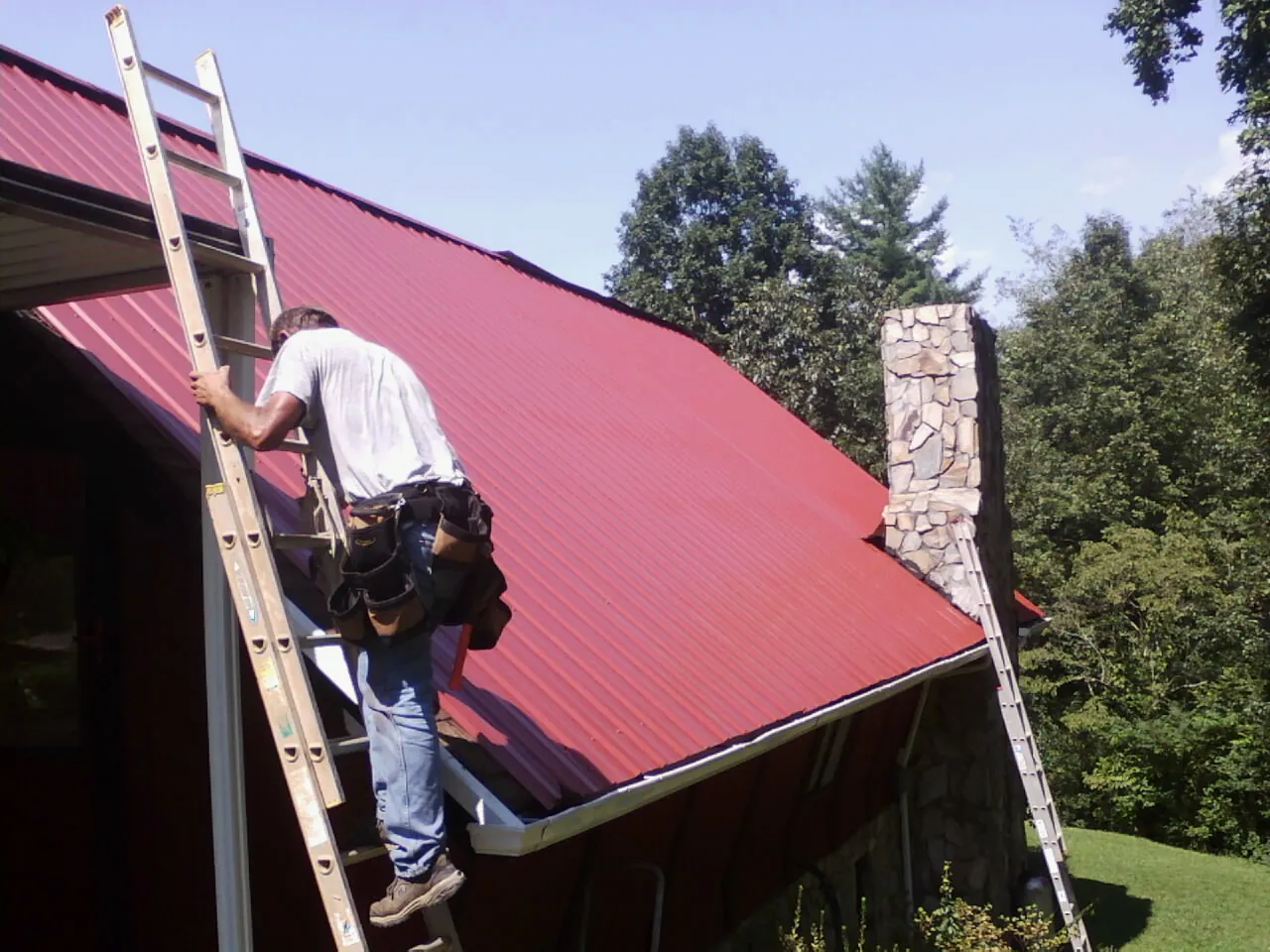 Roofer on a ladder beside a red metal roof and stone chimney — Soffit & Fascia Repair by Roofing Brothers in Cave Springs